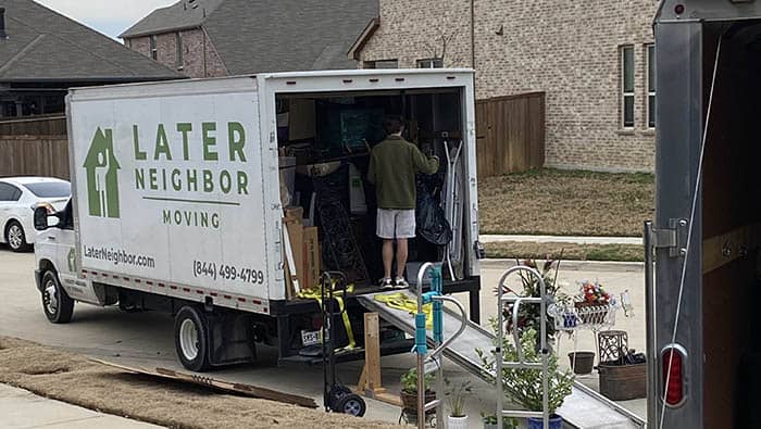 A white branded moving truck from Later Neighbor Moving is standing near a residential property almost full of residential furniture.